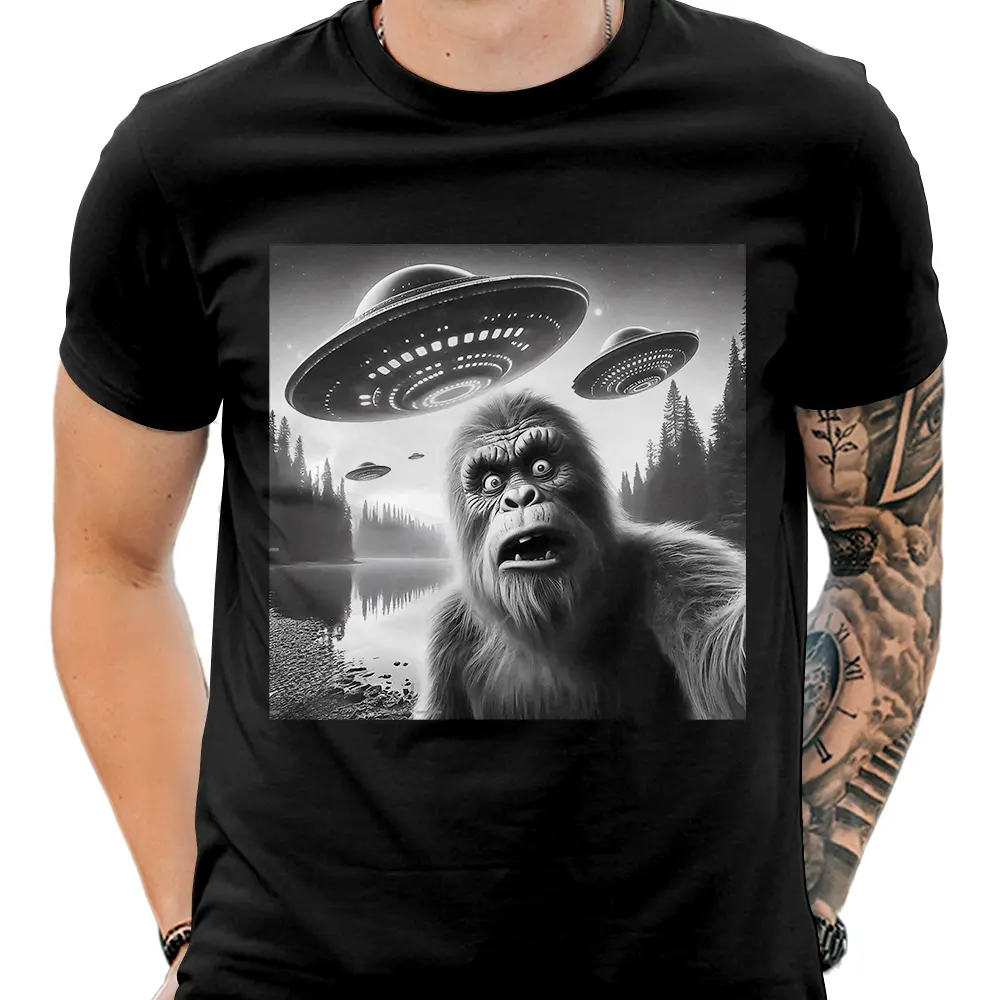 Black-and-white t-shirt design featuring a shocked Bigfoot with UFOs hovering above a forested lakeshore